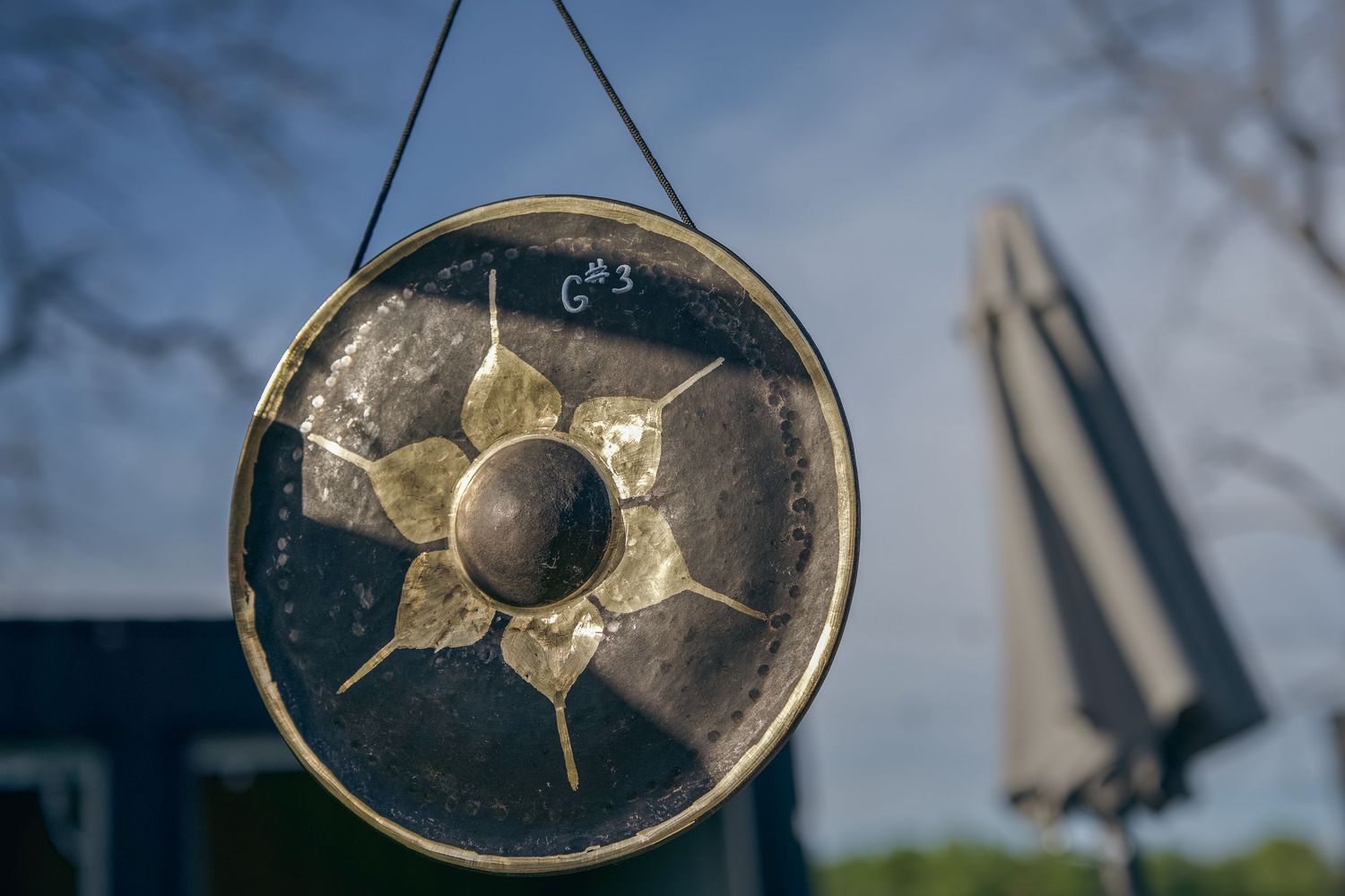 A decorative metal gong with a golden flower center, labeled G3, hanging outdoors against a blurred blue sky.