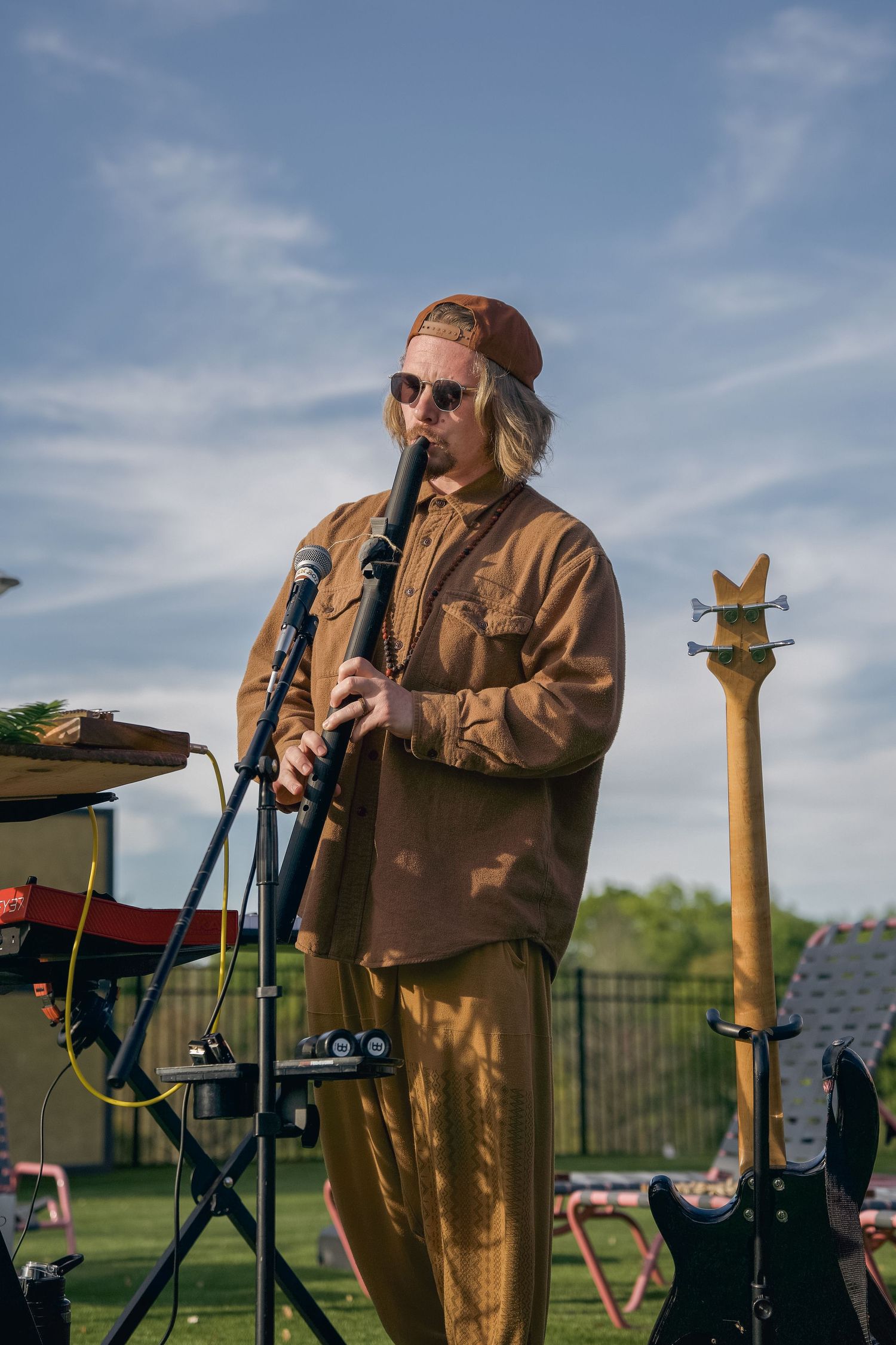 Musician in brown outfit plays clarinet outdoors at a festival, with guitars and drums visible on a sunny day.