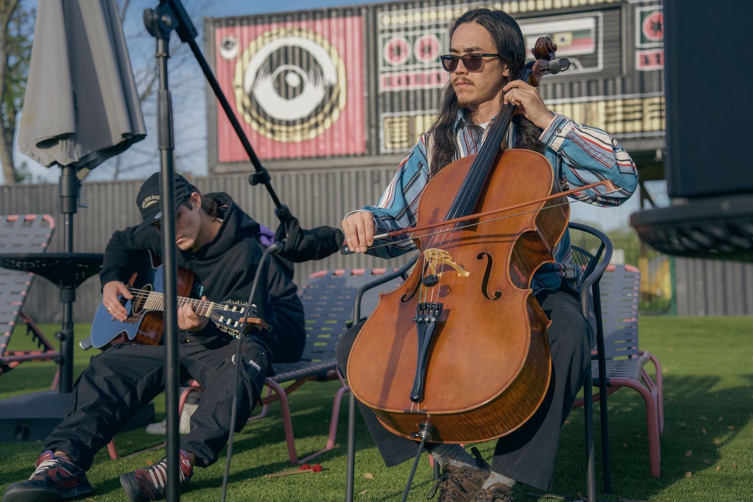 Two musicians perform outdoors, one playing cello and one guitar, at a festival stage.