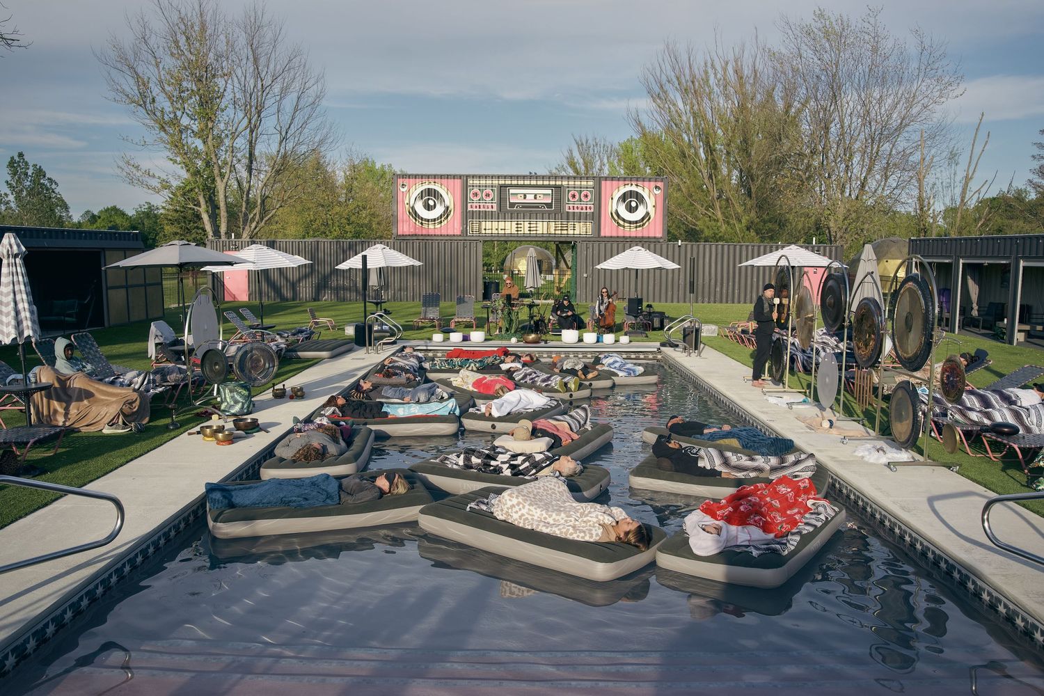 Outdoor pool party with floating loungers, umbrellas, and decorative backdrop in a landscaped garden setting.