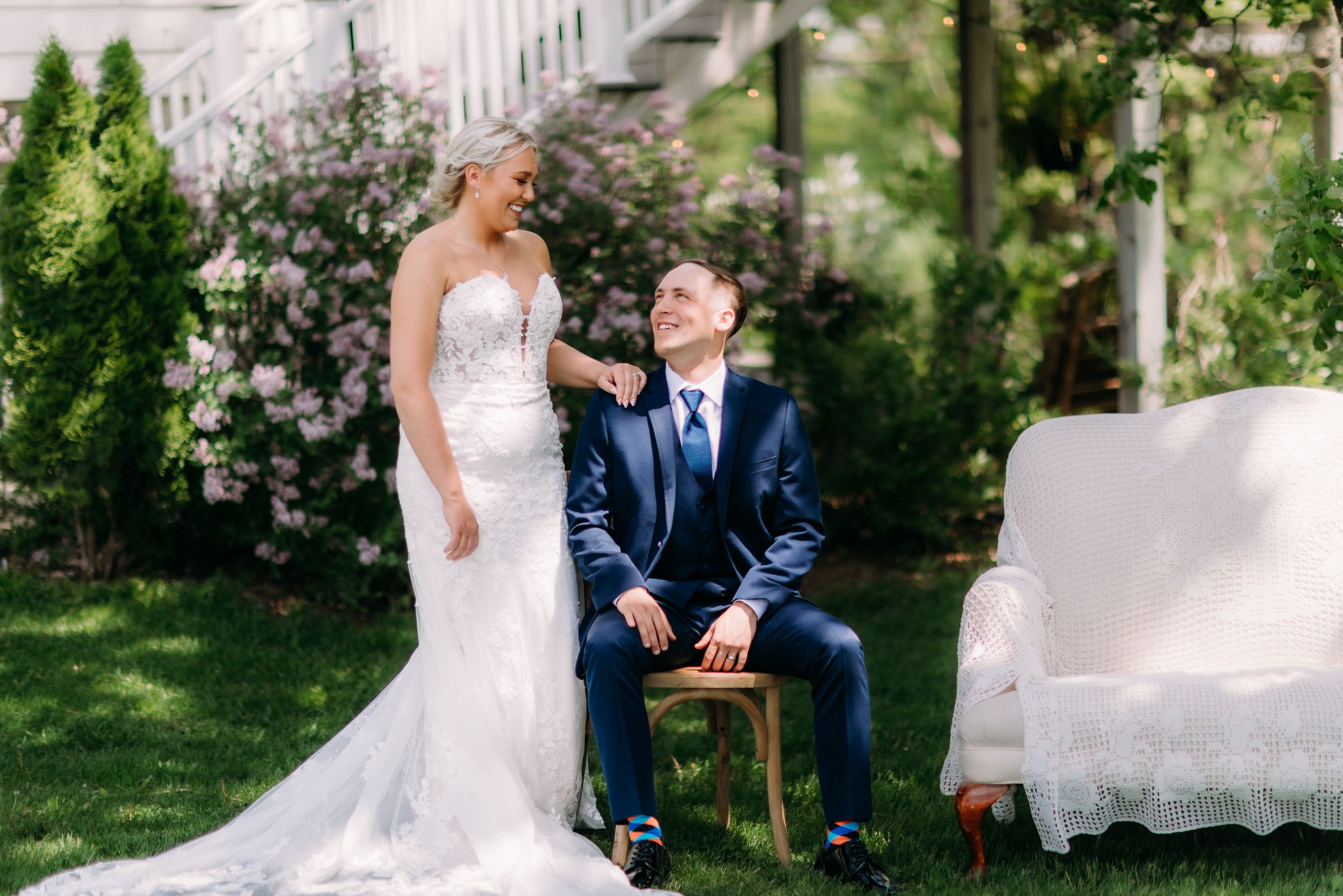 Bride in lace gown and groom in navy suit share a joyful moment outdoors on a sunny wedding day.