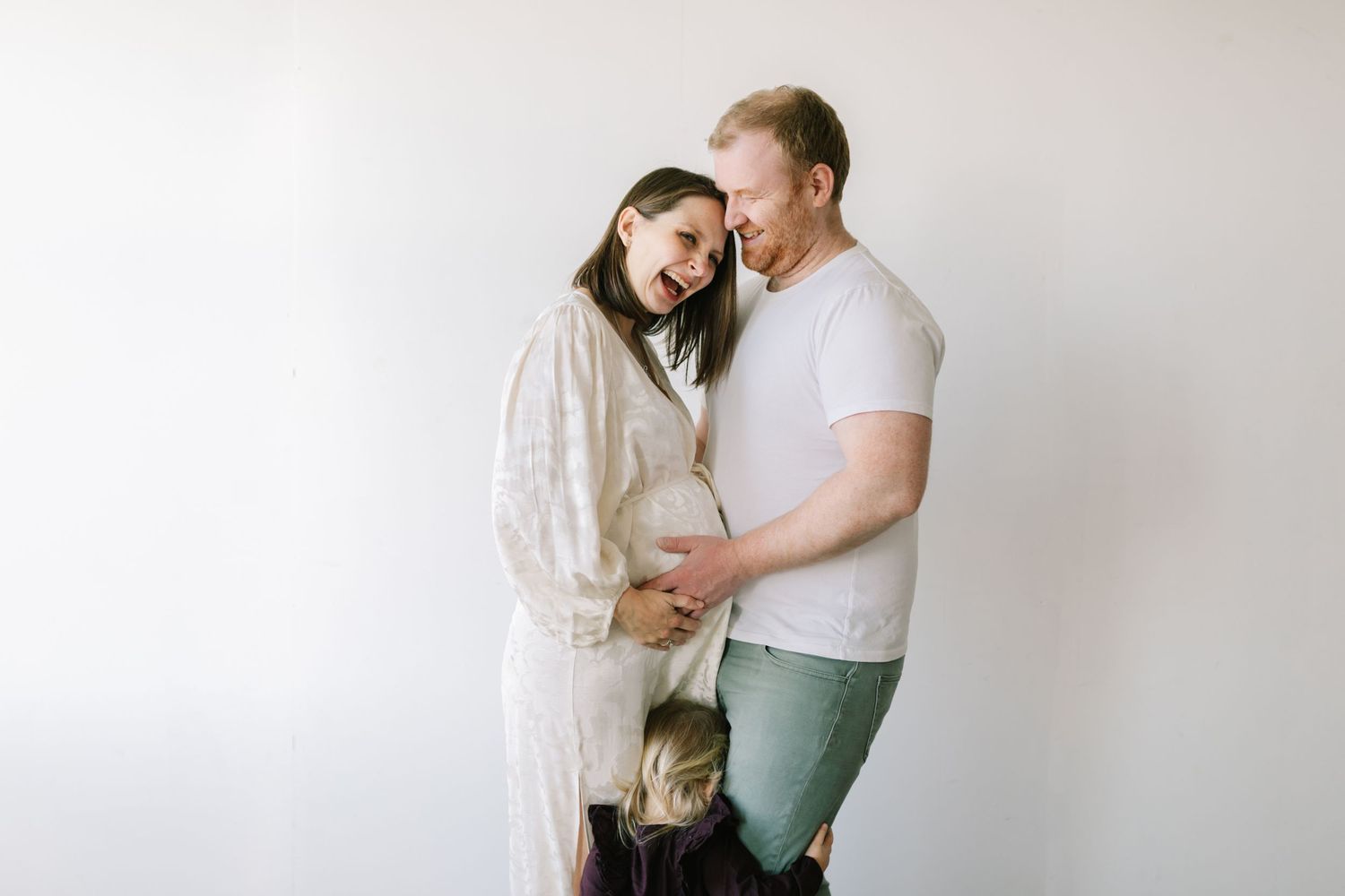 Maternity sessions, couple smiling together while cradling baby bump with a toddler standing between them.