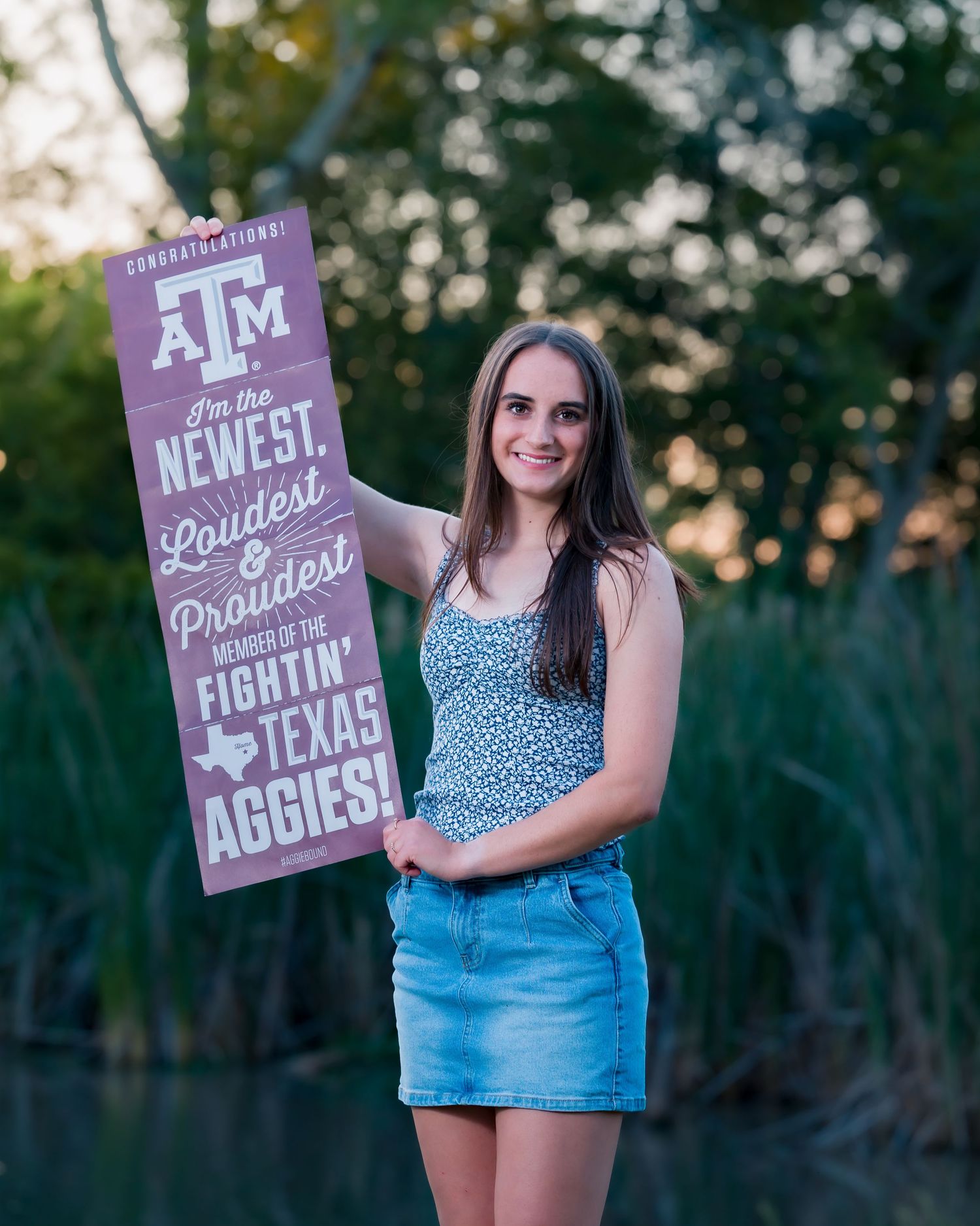 Young woman proudly holding a Texas A&M Fightin Aggies congratulations sign outdoors near a lake.