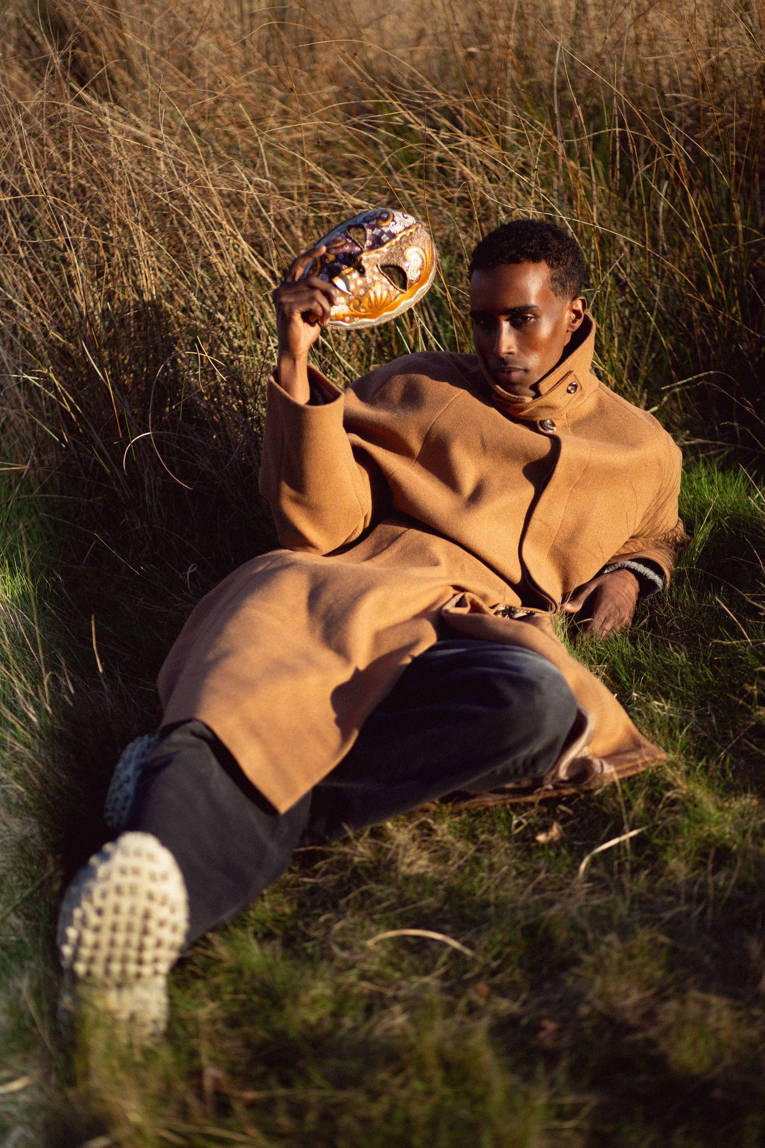 Man in brown hoodie sitting outdoors holding a decorative golden mask among tall grass in natural light.