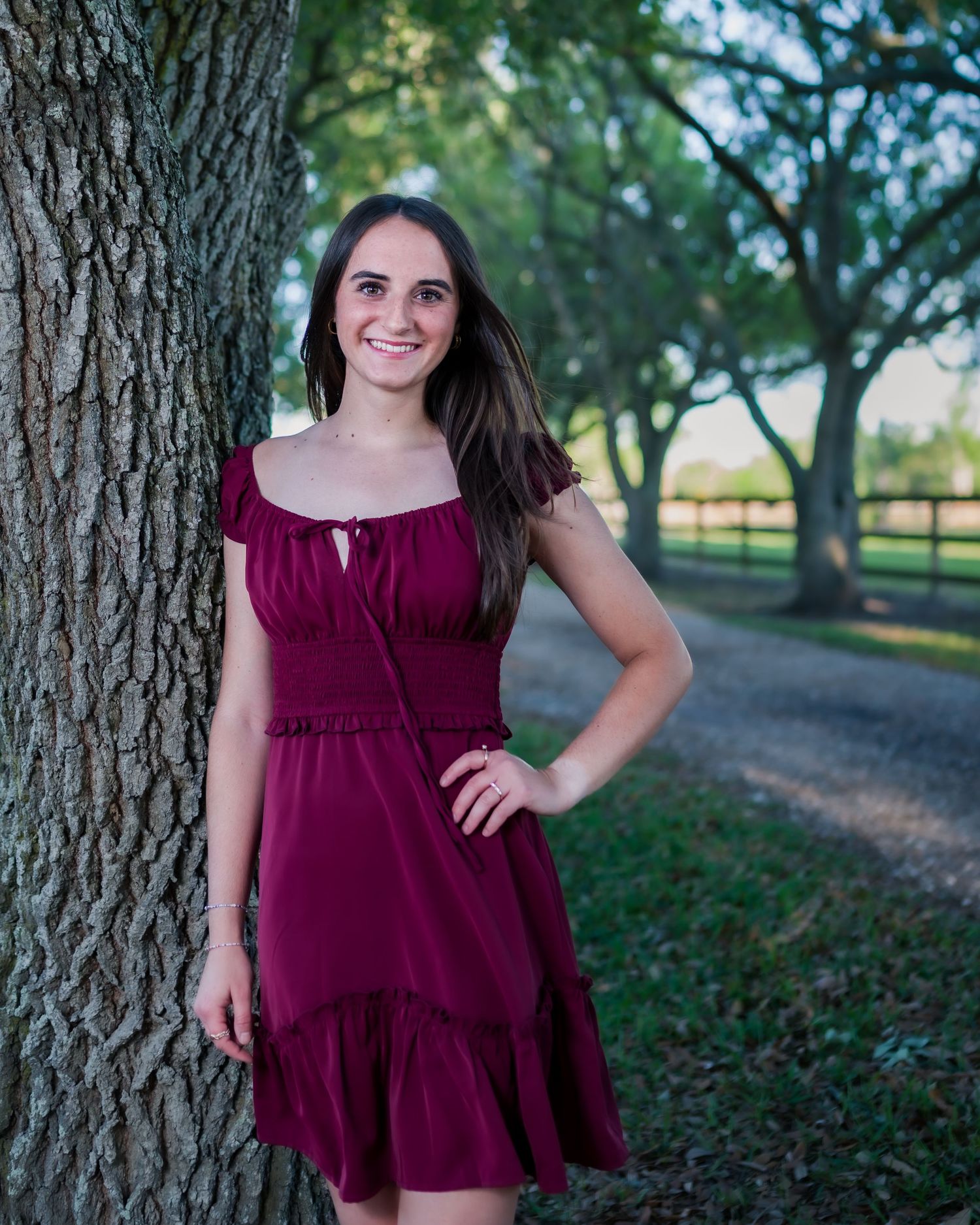Young woman in burgundy dress smiling beside a tree in a scenic outdoor pathway with lush greenery.