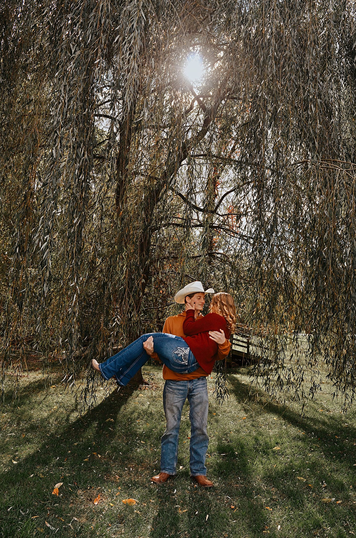 A young couple holds hands and walks through a lush green park surrounded by colorful autumn trees.