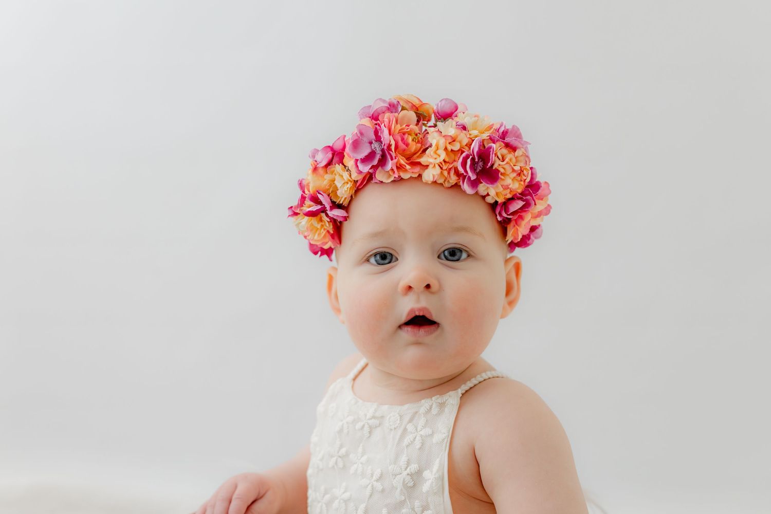 Adorable baby girl wearing a colorful pink and orange floral crown, dressed in white lace, against a light background.