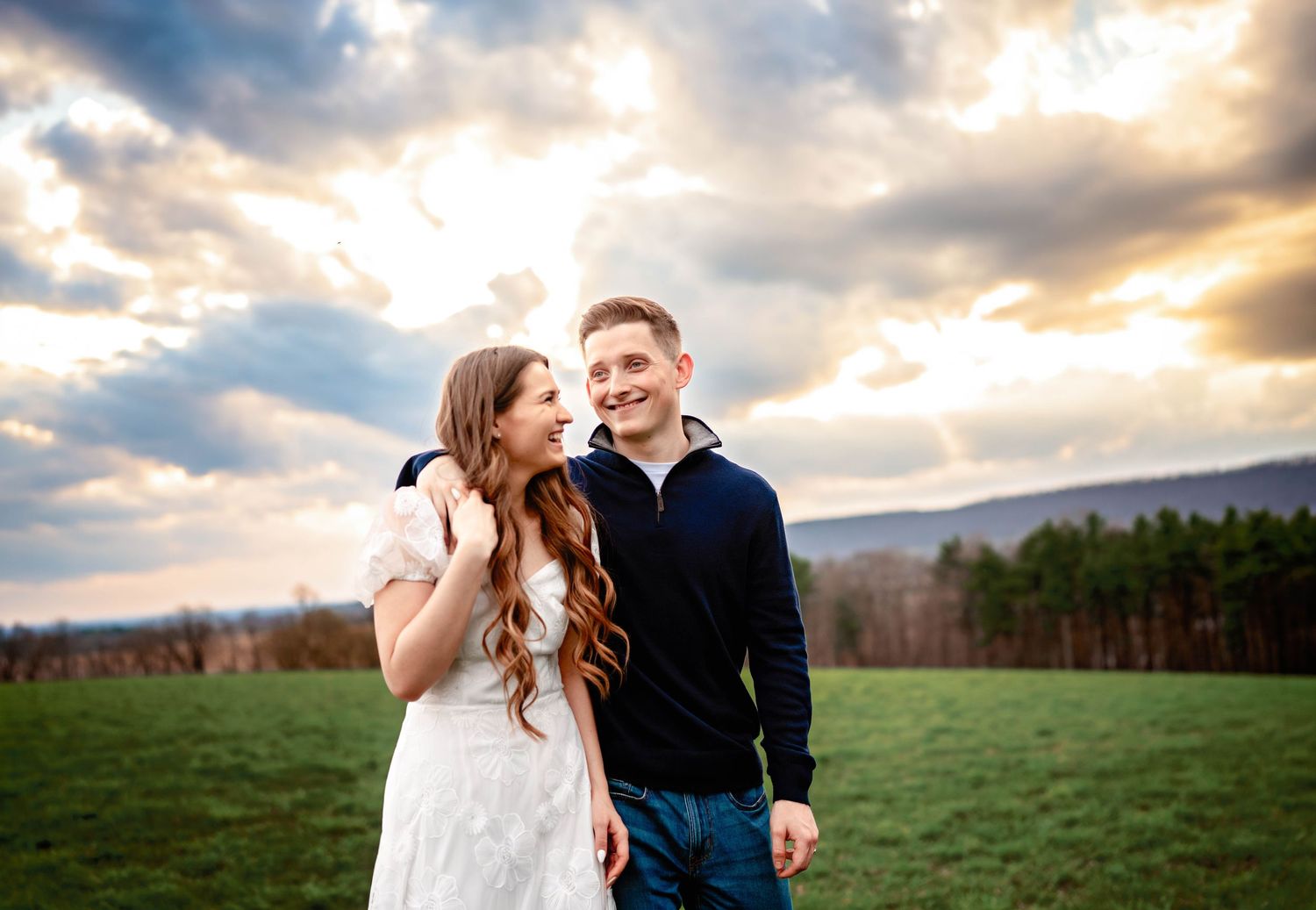 A smiling young couple poses outdoors on a green field with a dramatic cloudy sky at sunset.