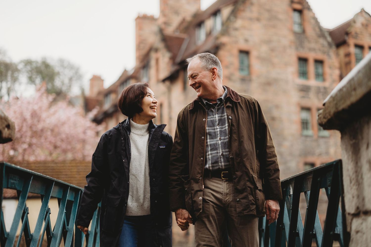 Smiling older couple holding hands on a bridge in a charming historic town with cherry blossoms in the background.