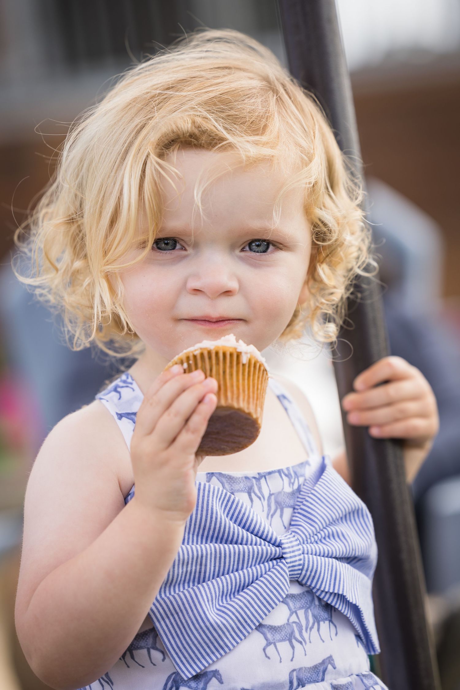 Cute toddler girl with curly blonde hair eating a chocolate cupcake outdoors in a blue striped dress.