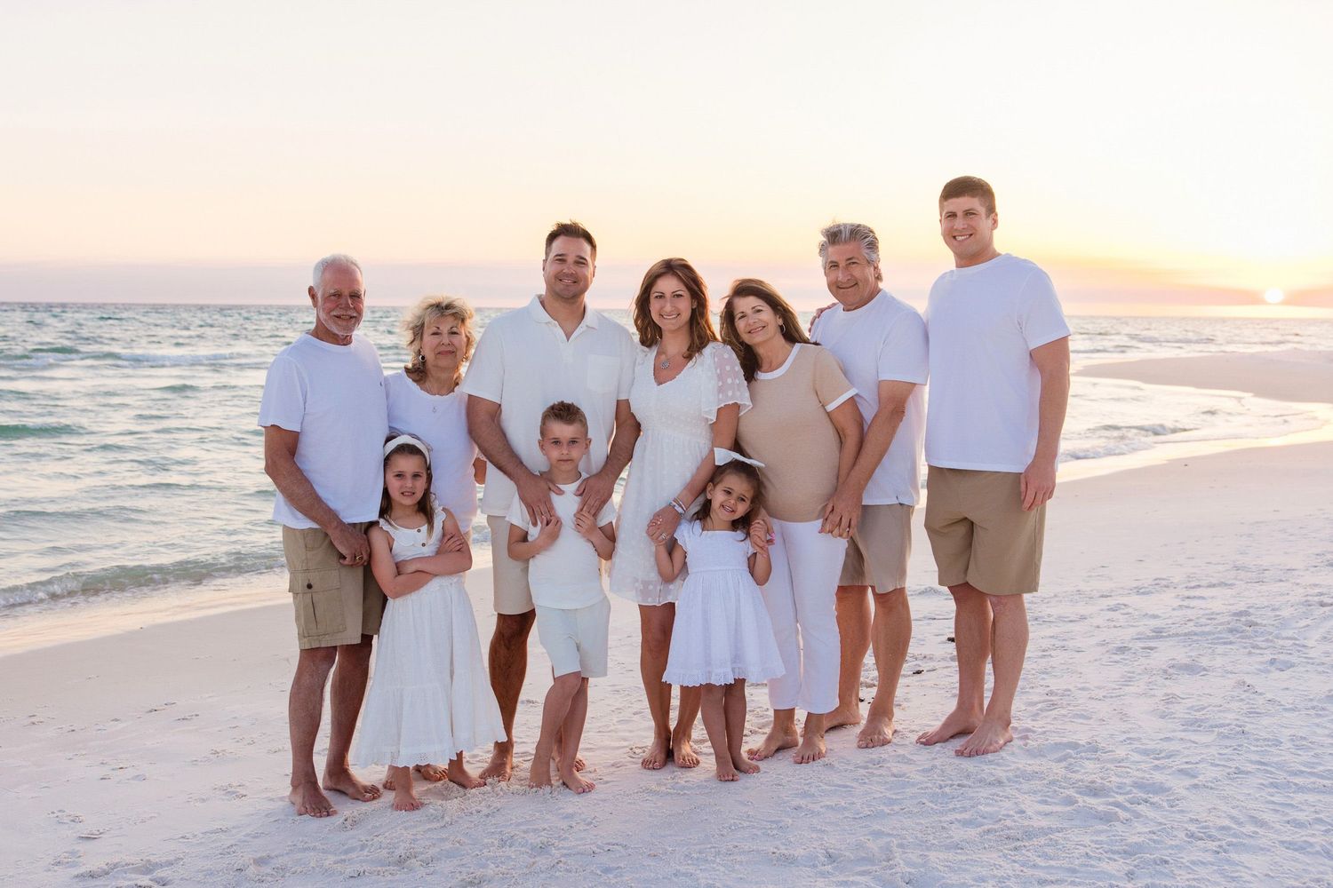Multi-generational family in white and tan outfits posing together on a sandy beach at sunset.