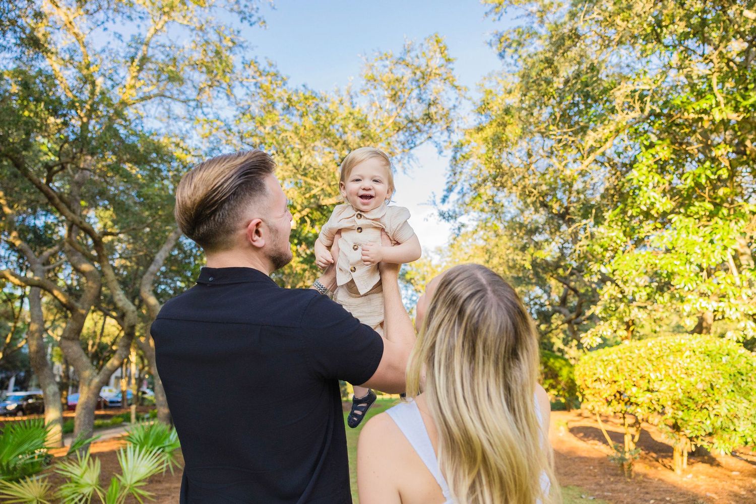 A young family lifting their laughing baby in the air outdoors among lush green trees on a sunny day.