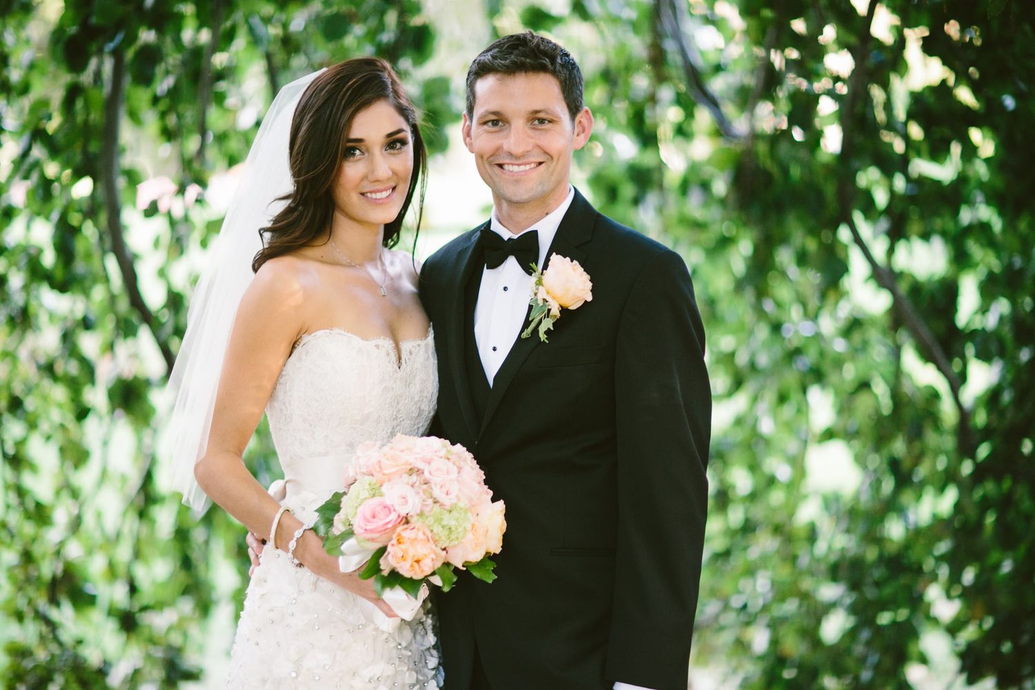 Newlyweds pose together outdoors with pink bouquet against green foliage.