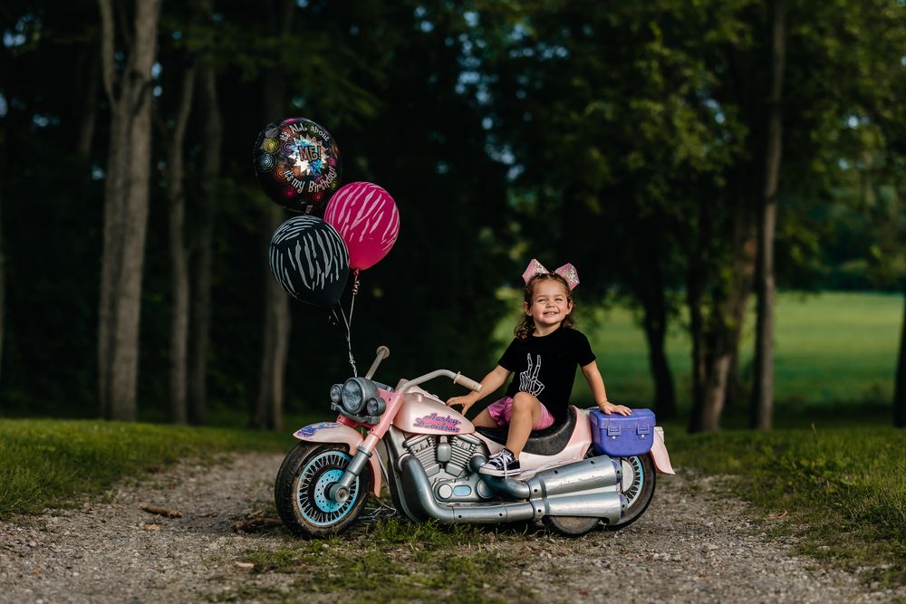 Young girl sitting on pink toy motorcycle with colorful balloons in a tree-lined outdoor setting.