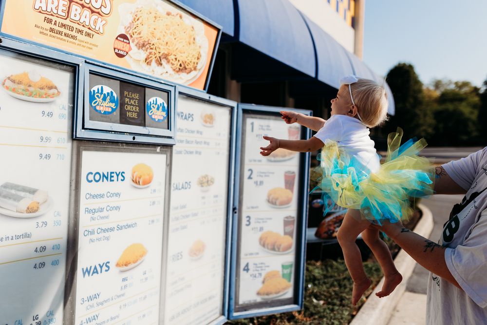 Baby girl in teal tutu being held up to view menu at Skyline Chili Coney restaurant outdoor ordering board.