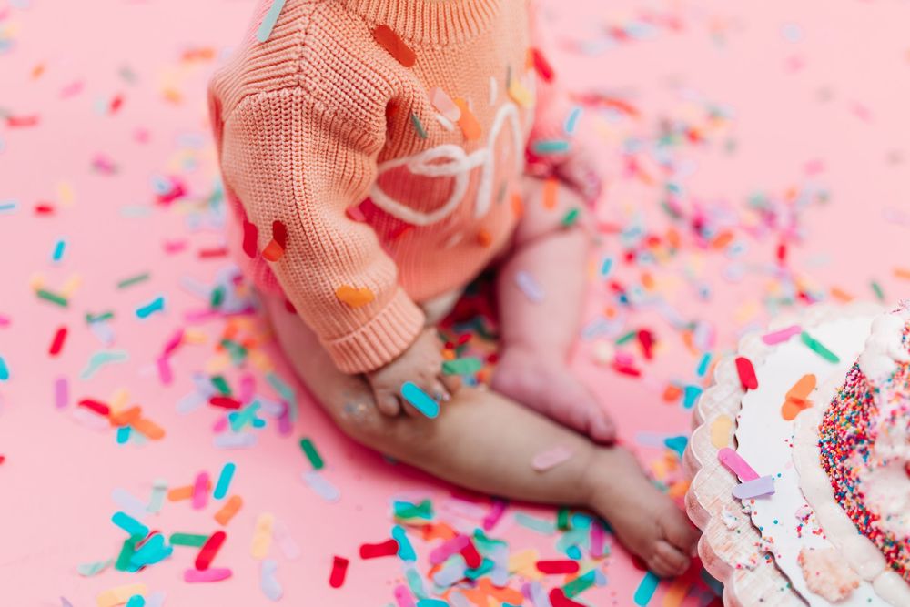 Close-up of baby girl's hand covered in colorful sprinkles beside smash cake on pink background.