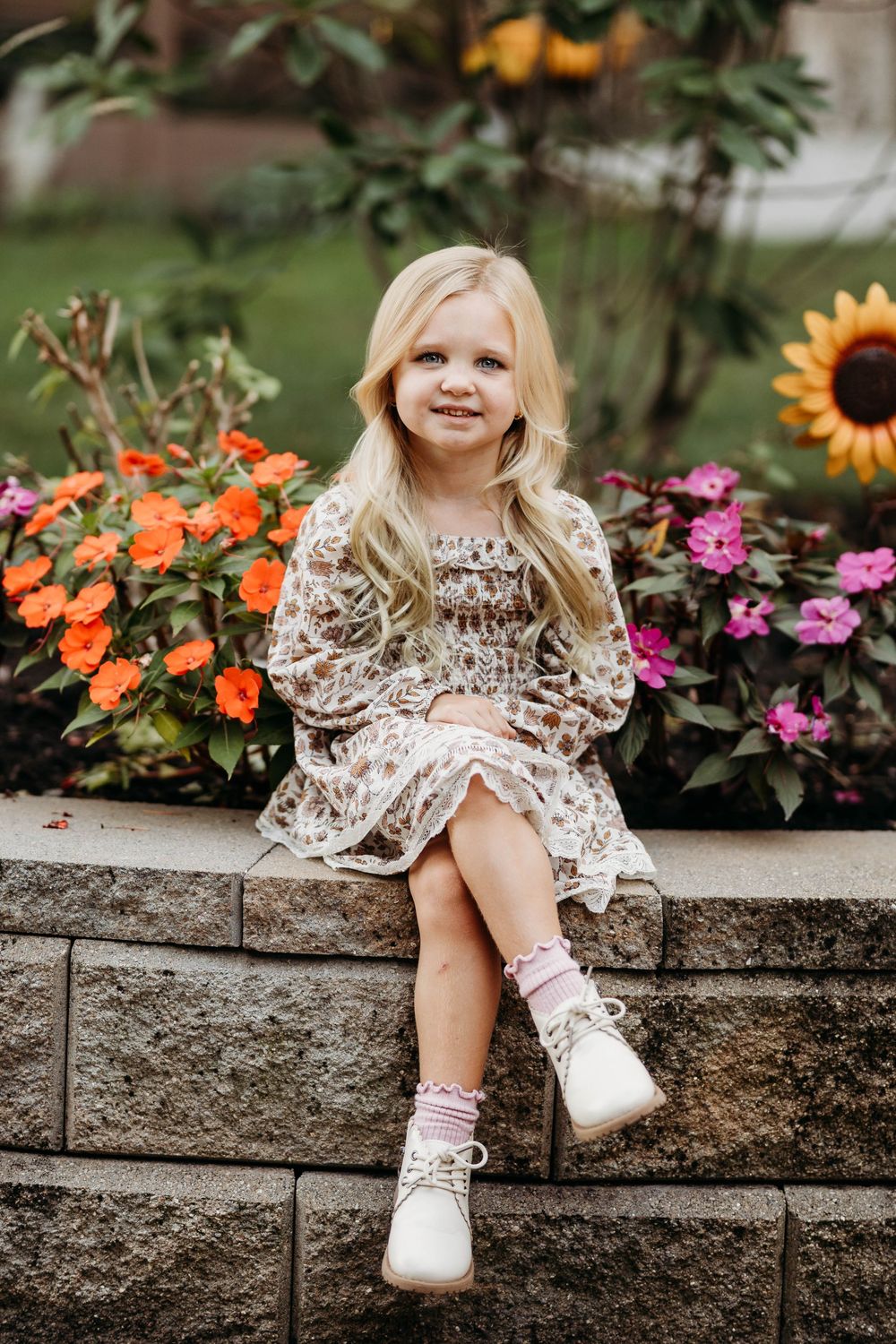 Cute blonde girl in floral dress sitting on stone wall surrounded by colorful garden flowers.