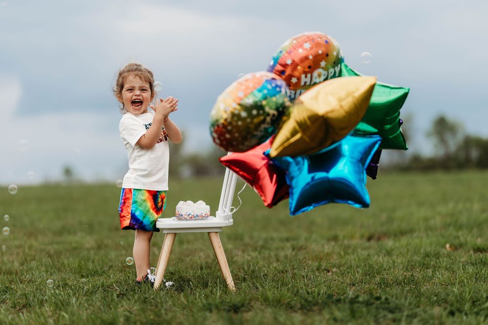 Excited toddler clapping hands beside small birthday cake and colorful foil balloons in a field.