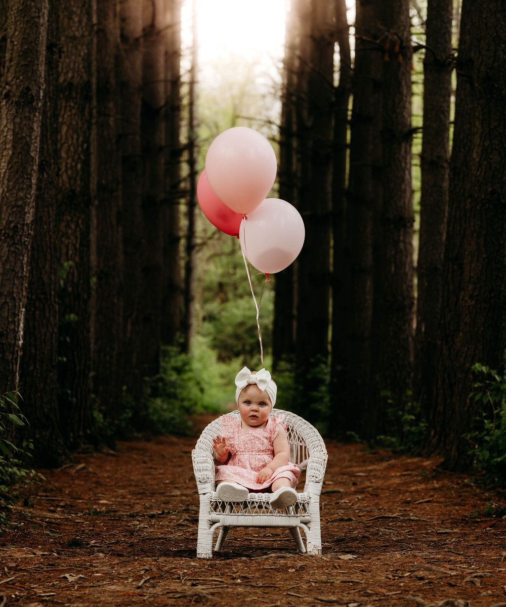 Baby girl in pink dress with white bow sits in wicker chair holding pink balloons in forest path.