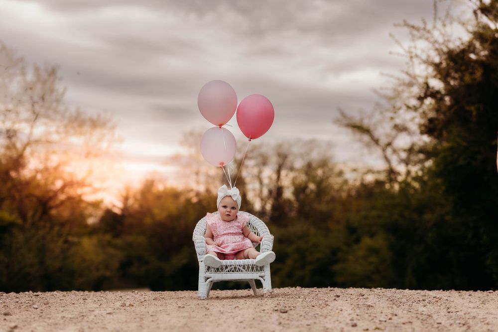 Baby girl in pink outfit sits in white wicker chair with pink balloons at sunset outdoors.
