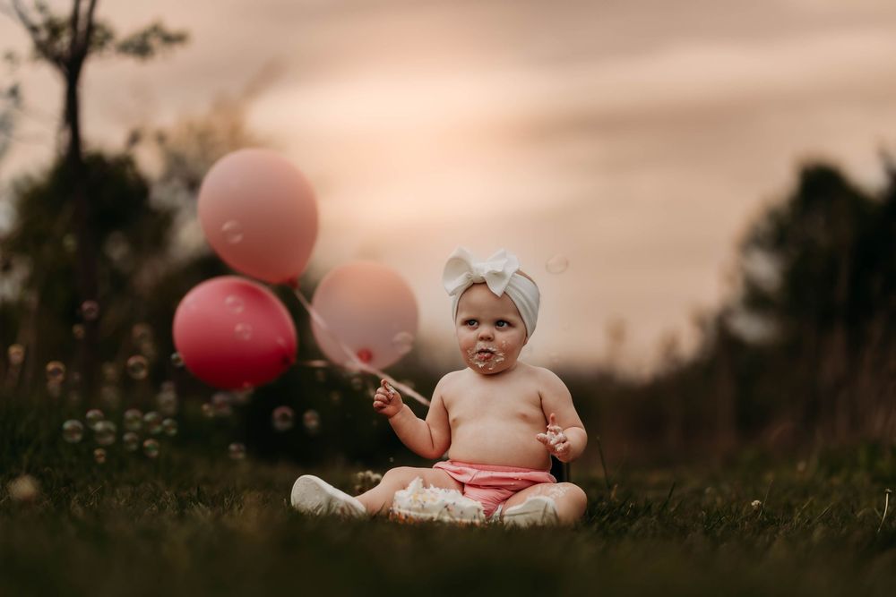 Baby girl in pink bloomers with white bow headband holds pink balloons while sitting in grass at sunset.