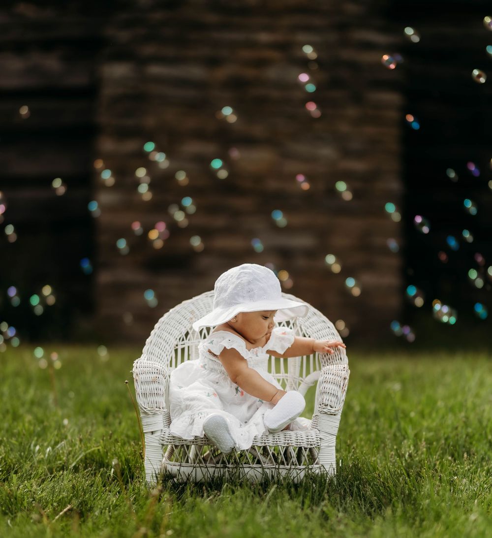 Baby in white hat sits in wicker chair surrounded by colorful floating bubbles outdoors.