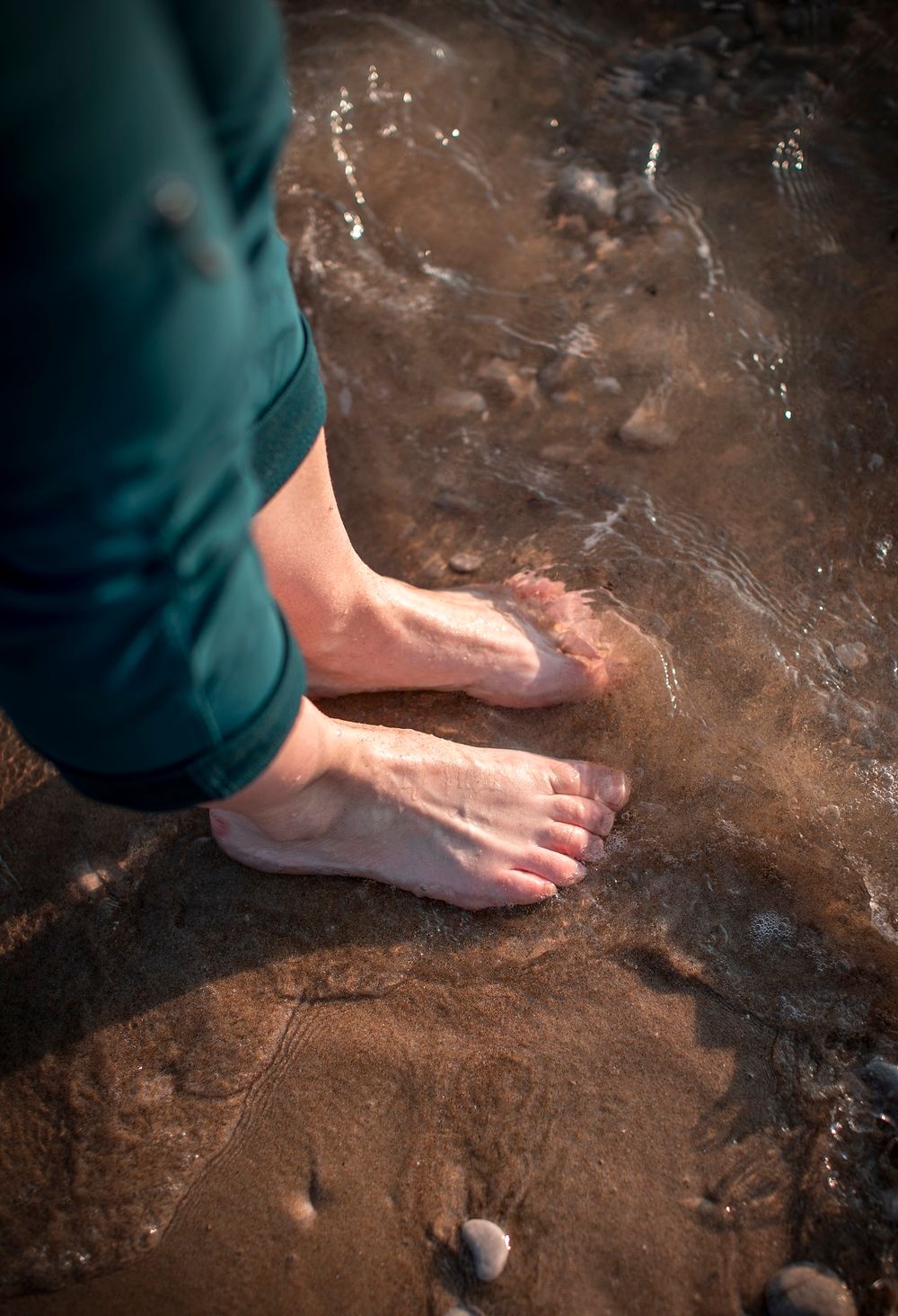Bare feet standing on wet sandy riverbed with small pebbles and flowing shallow water.