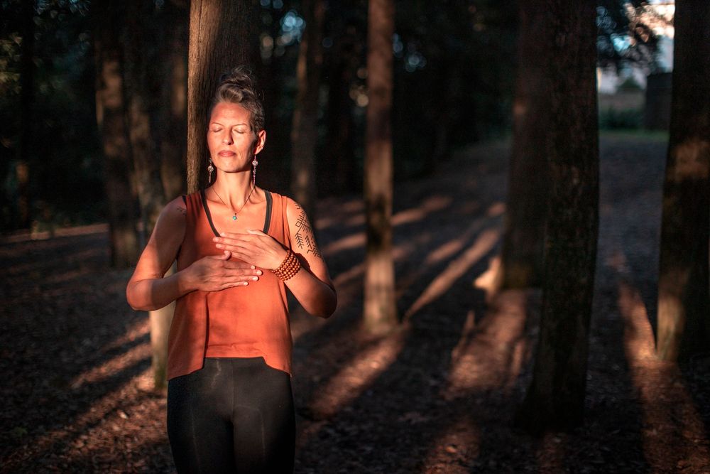 Woman meditating in forest at dusk, hands on chest, eyes closed, wearing orange top among tall trees.