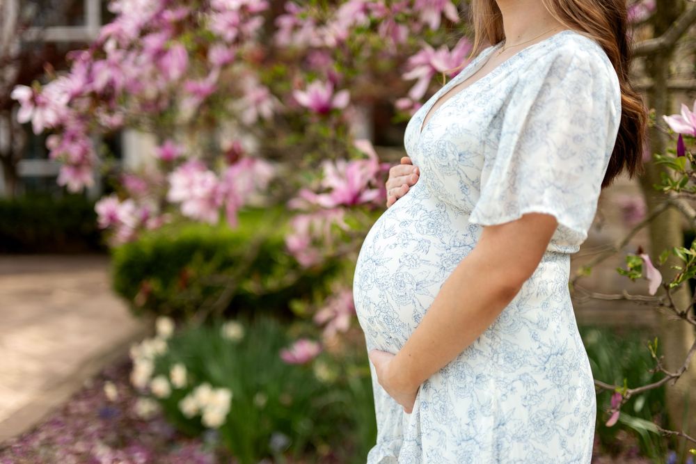 Close-up of pregnant woman in blue floral dress cradling baby bump surrounded by spring blooms.