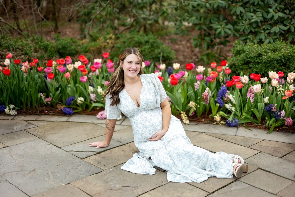 Smiling pregnant woman in white floral dress sitting on stone patio beside colorful spring tulip garden.