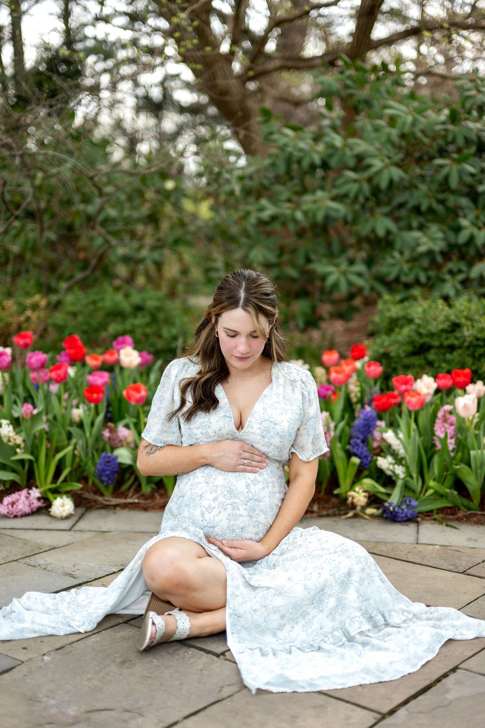 Pregnant woman in white floral dress sitting on stone patio gazing at bump near vibrant tulip garden.