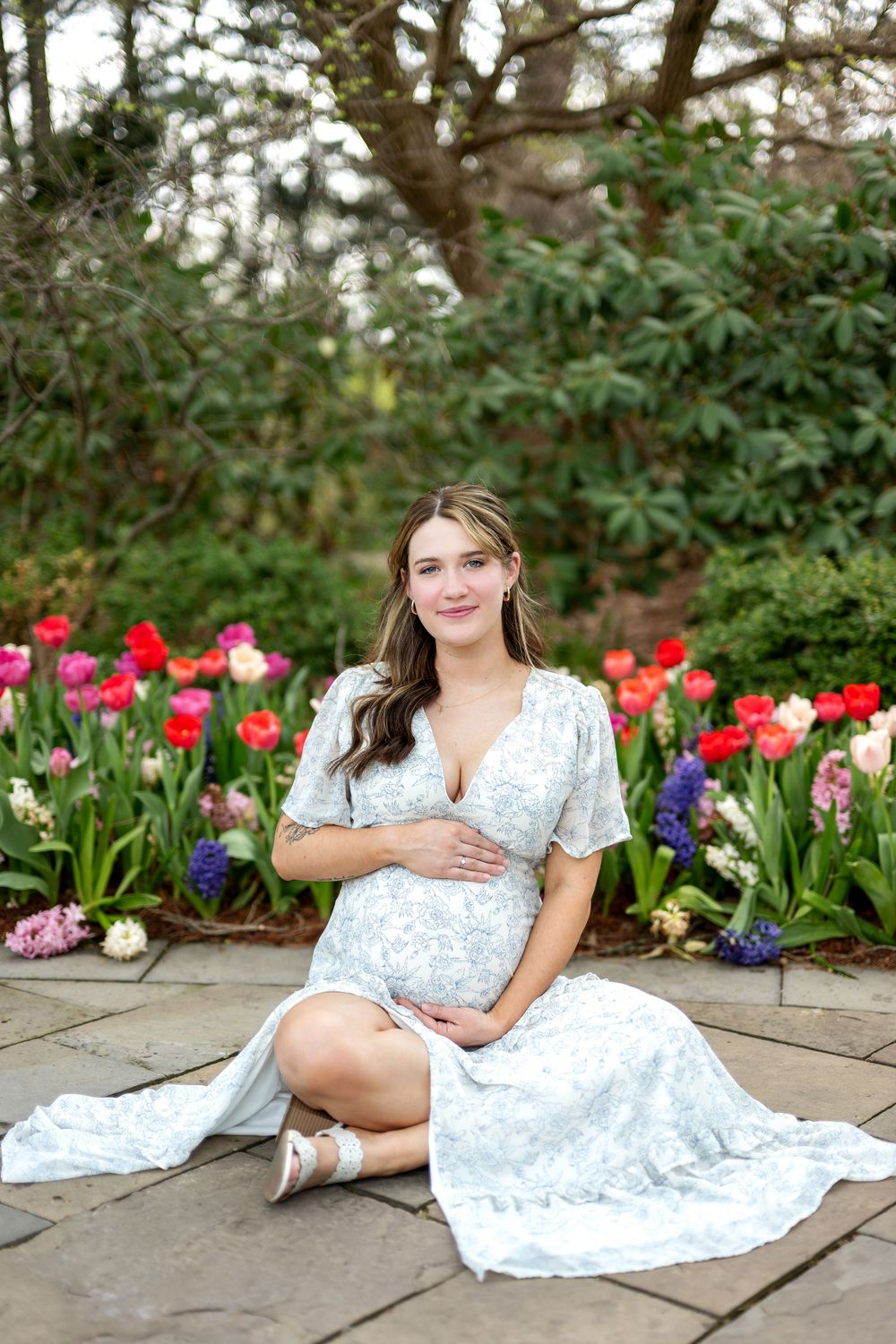 Pregnant woman in white floral dress sitting gracefully on stone patio in front of colorful tulip garden.