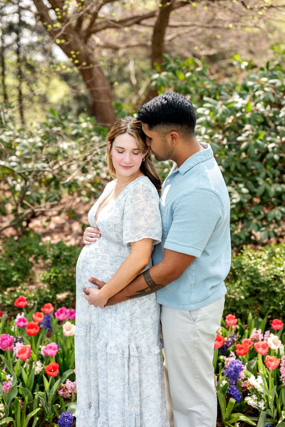 Couple embracing among spring tulips, man nuzzling pregnant woman in light blue floral maternity dress.