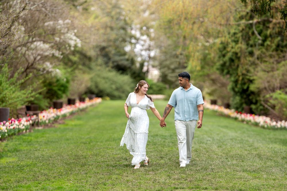 Happy pregnant couple walks hand-in-hand through a vibrant tulip garden path in spring bloom.