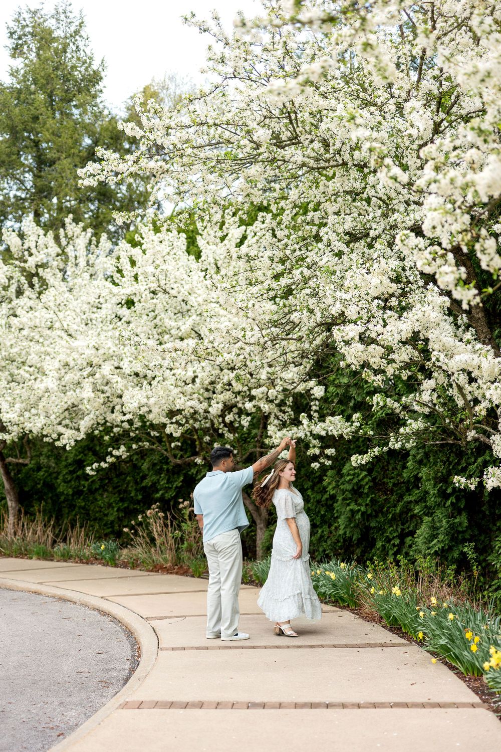 Man gently placing flowers in pregnant woman's hair near white blooming trees on a garden path.
