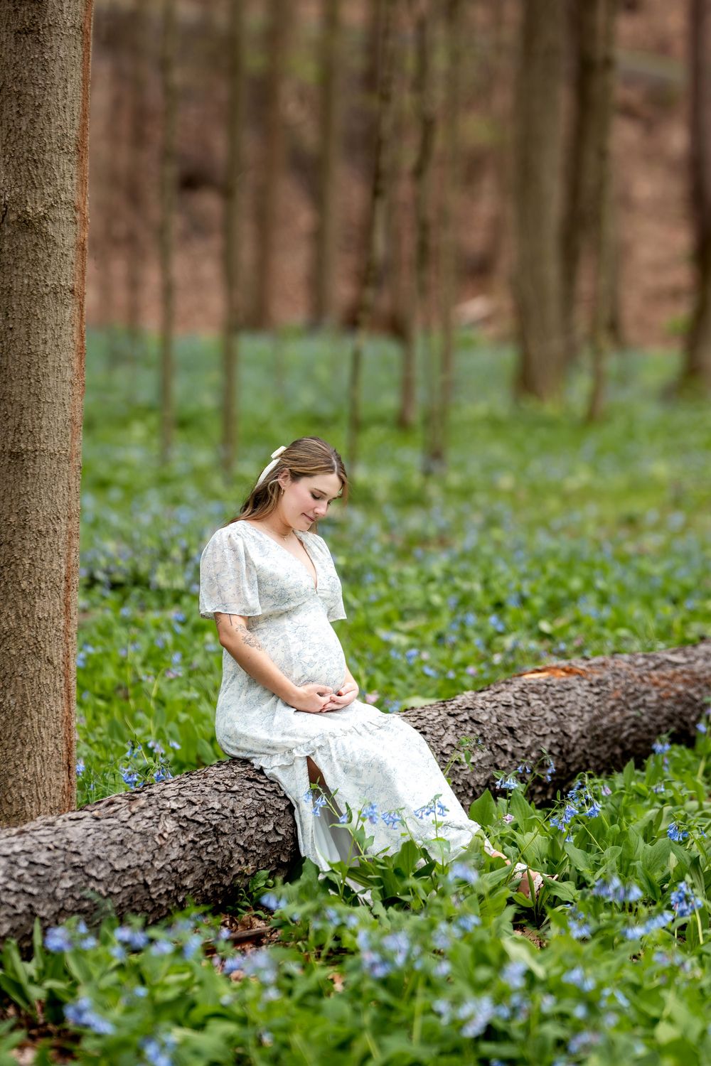 Pregnant woman in white lace dress sitting on a fallen log in a bluebell woodland.