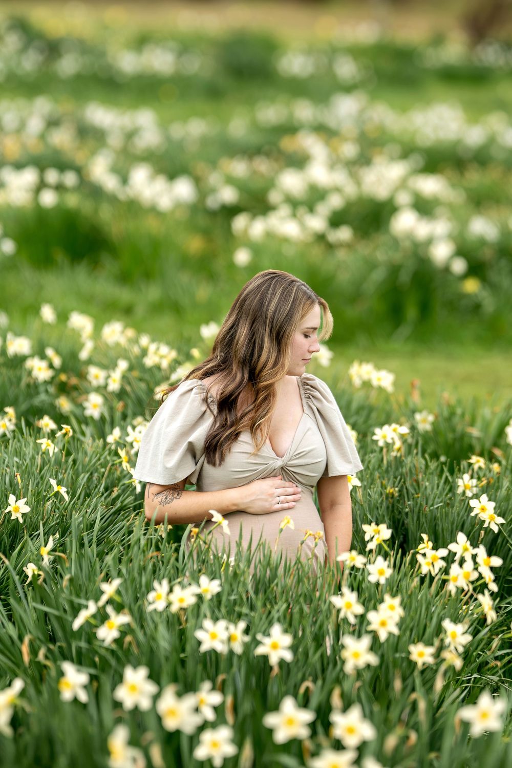 Pregnant woman in beige dress standing among yellow daffodils in a lush green field.