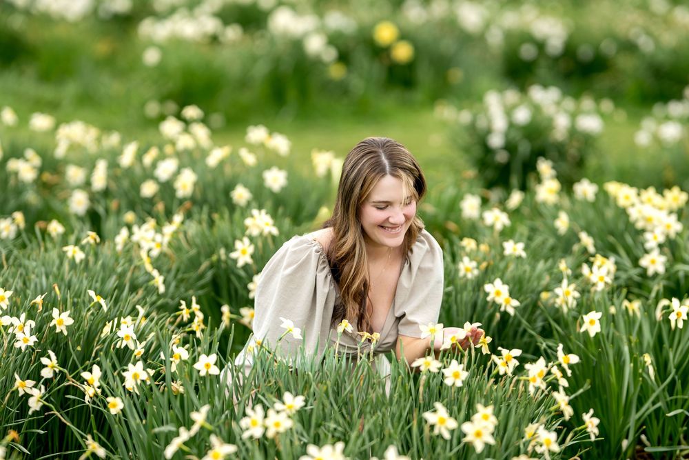 Woman smiling while sitting among blooming yellow daffodils in a green meadow.