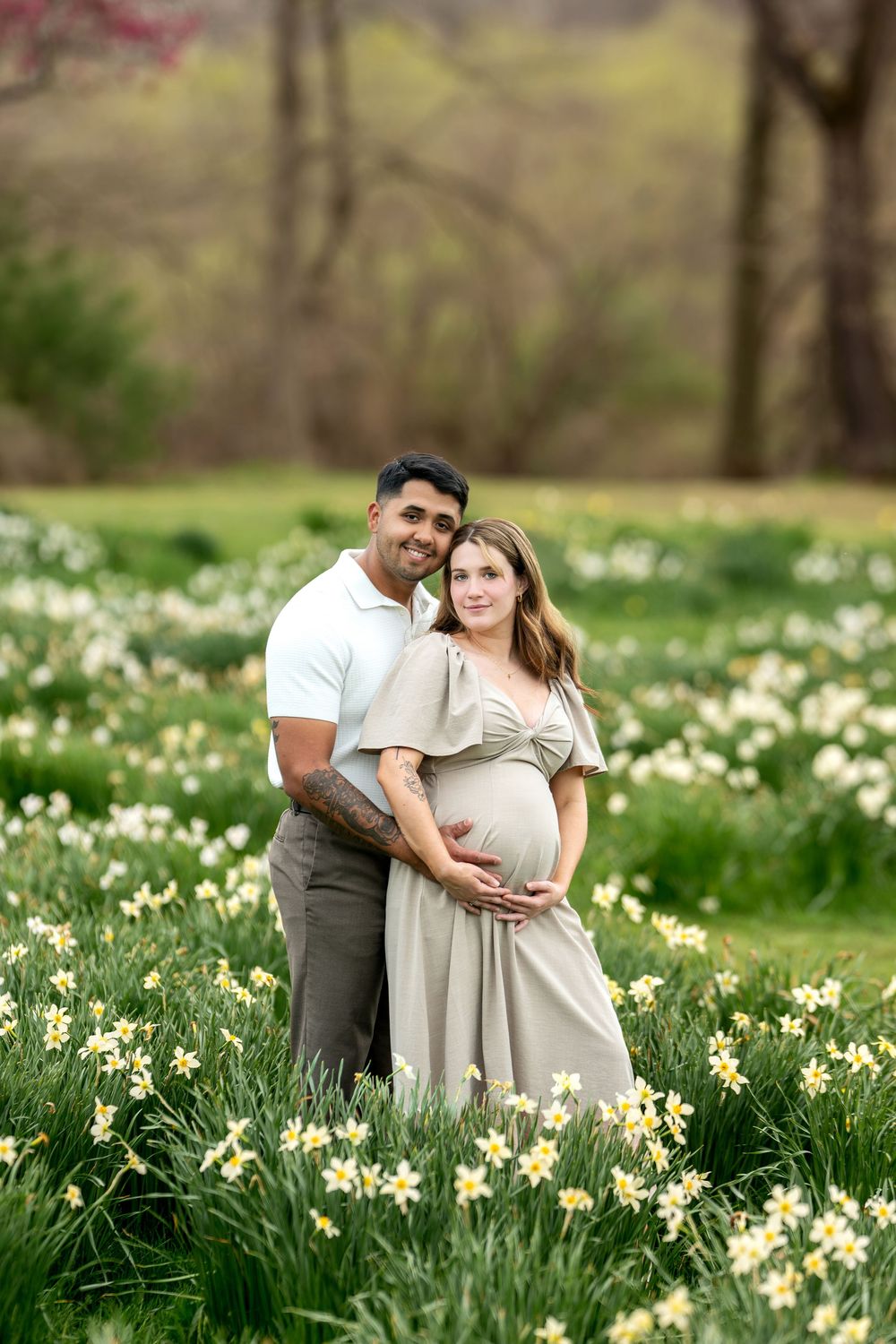 Couple embracing in a field of yellow daffodils, woman in beige maternity dress.