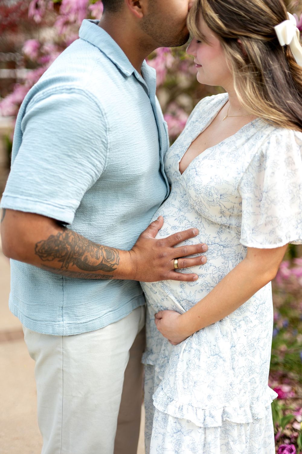 Man kissing pregnant partner while tenderly holding her baby bump outdoors near pink flowers.