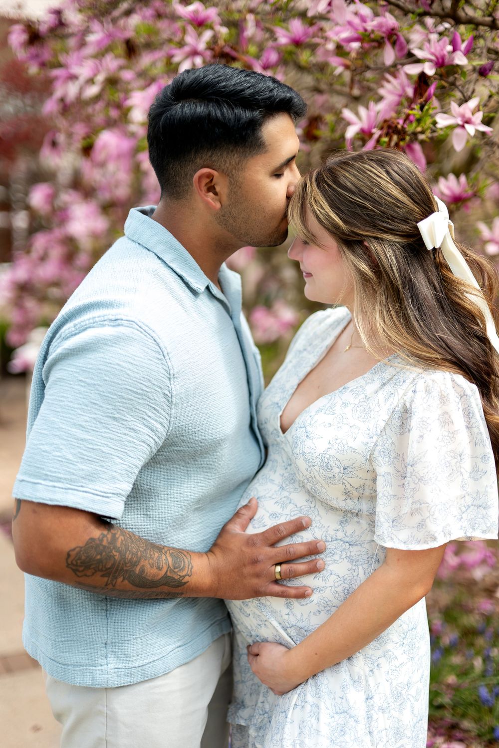 Man kissing pregnant woman on forehead while holding her baby bump near pink magnolia blossoms.