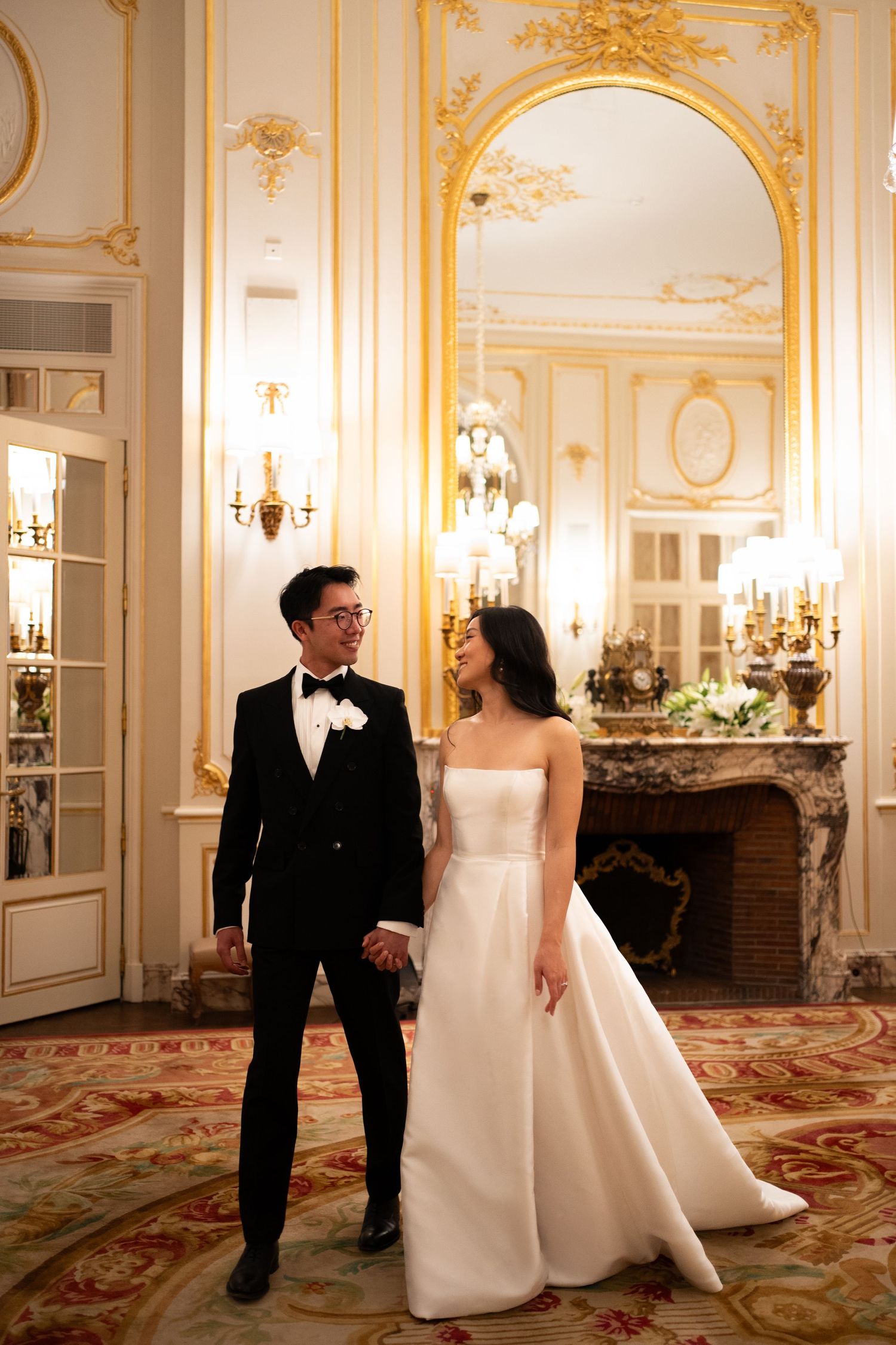 Bride and groom hold hands in an ornate gold ballroom with chandeliers and marble fireplace during wedding portrait.