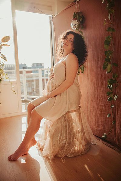 A pregnant woman poses in cream colored dress by window with warm natural lighting and trailing plants in Hoofddorp