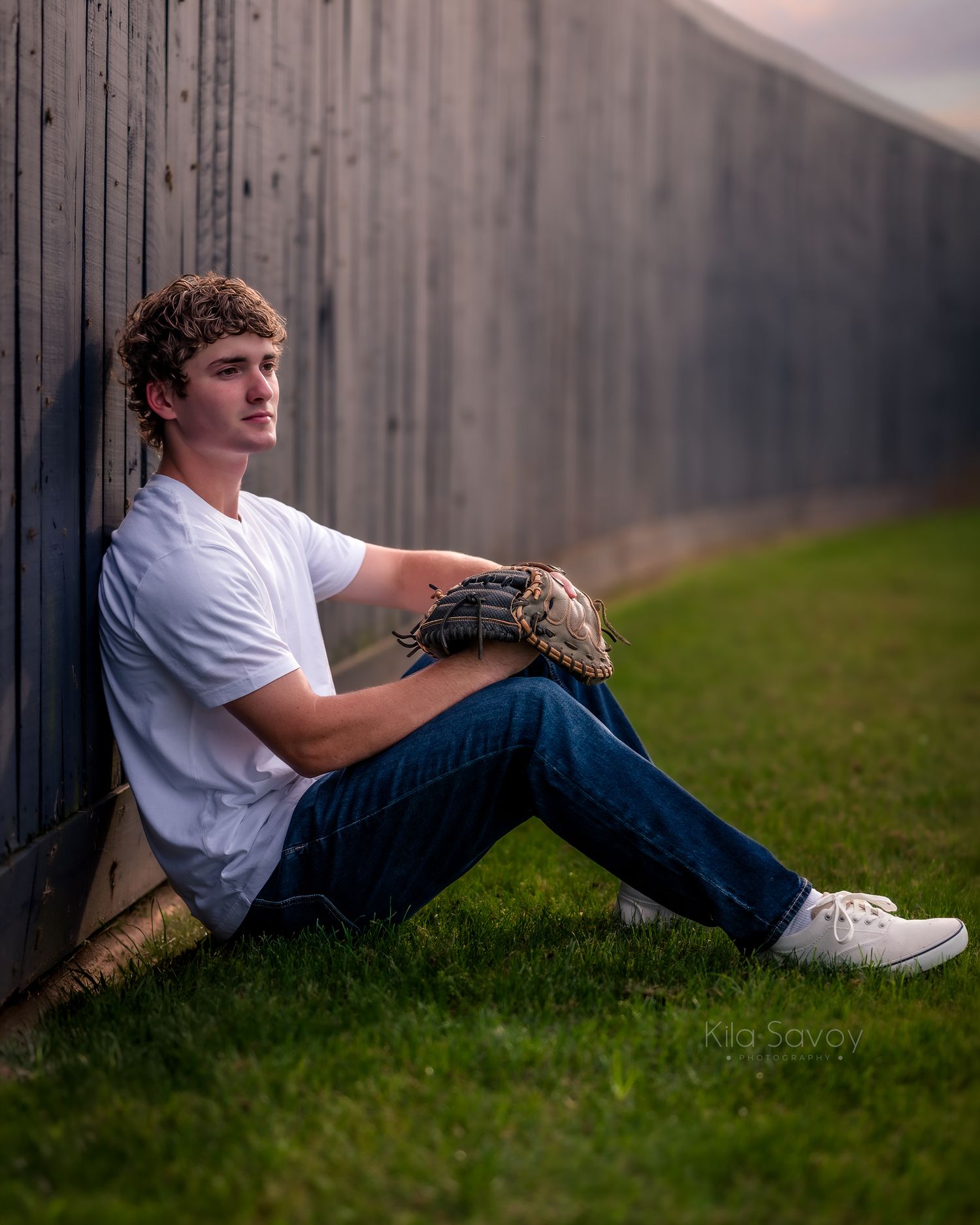 A young man in a white t-shirt and jeans sits on grass against a wooden fence holding a baseball glove.