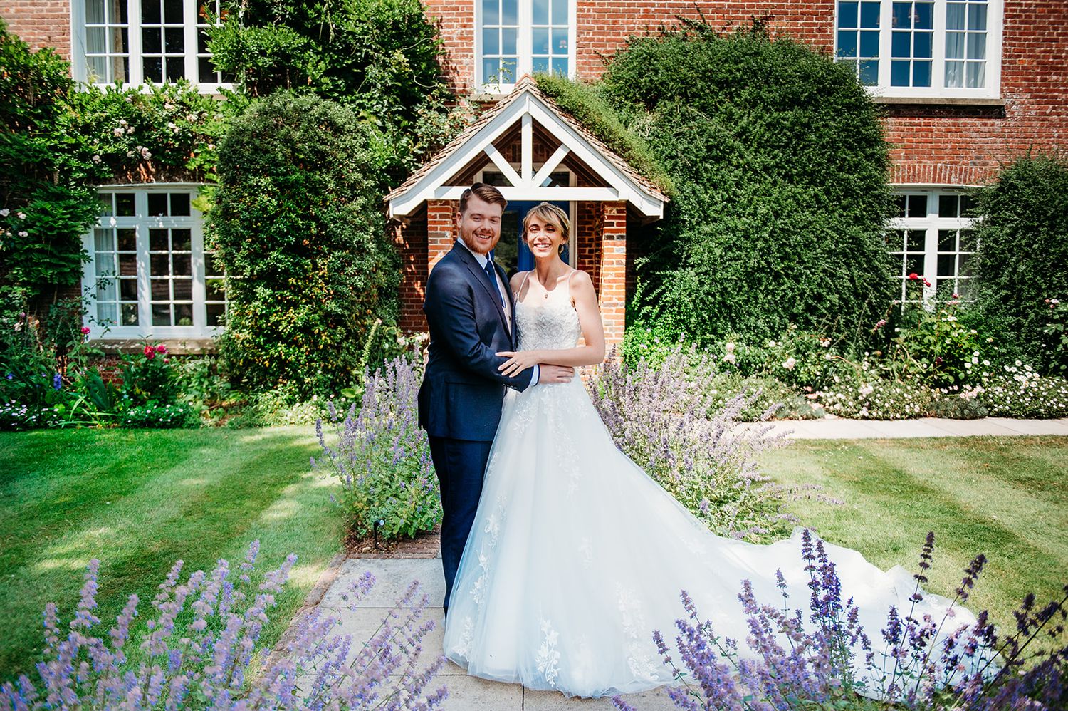 Bride and groom pose in a lush garden outside a charming brick manor house surrounded by lavender blooms.