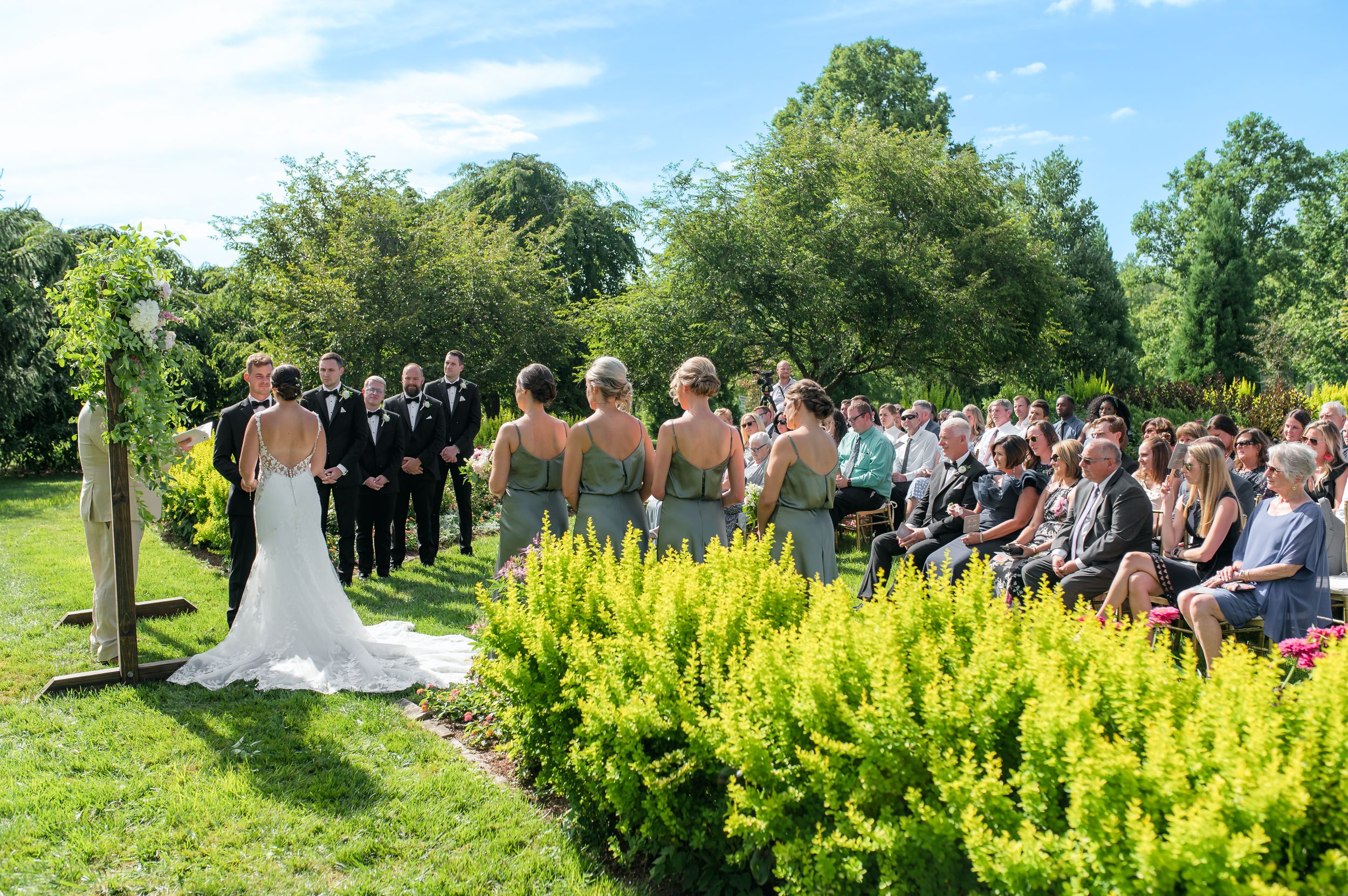 Outdoor wedding ceremony in a lush garden with bridesmaids, groomsmen, and seated guests on a sunny day.