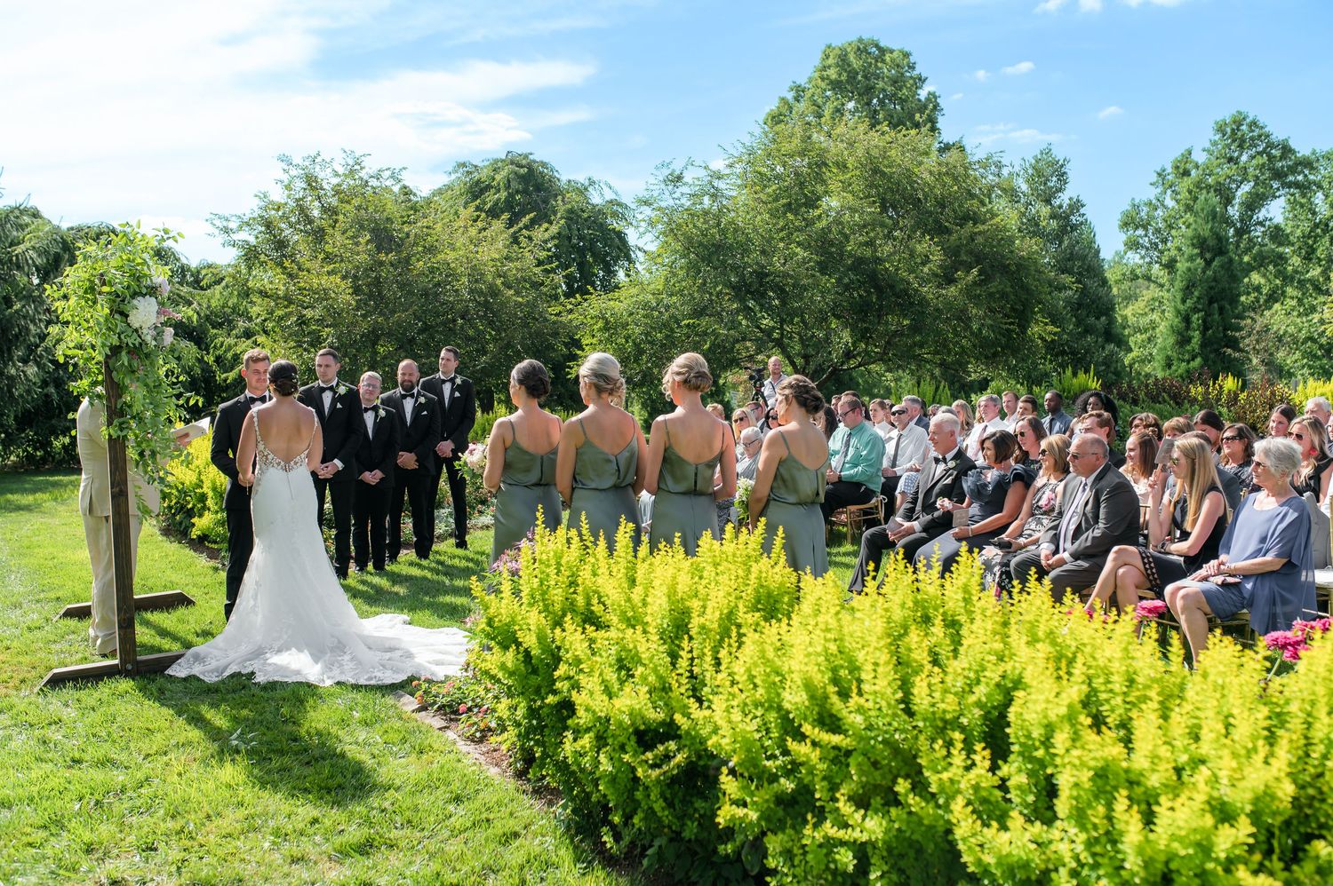 Outdoor wedding ceremony in a lush garden with bridesmaids, groomsmen, and seated guests on a sunny day.