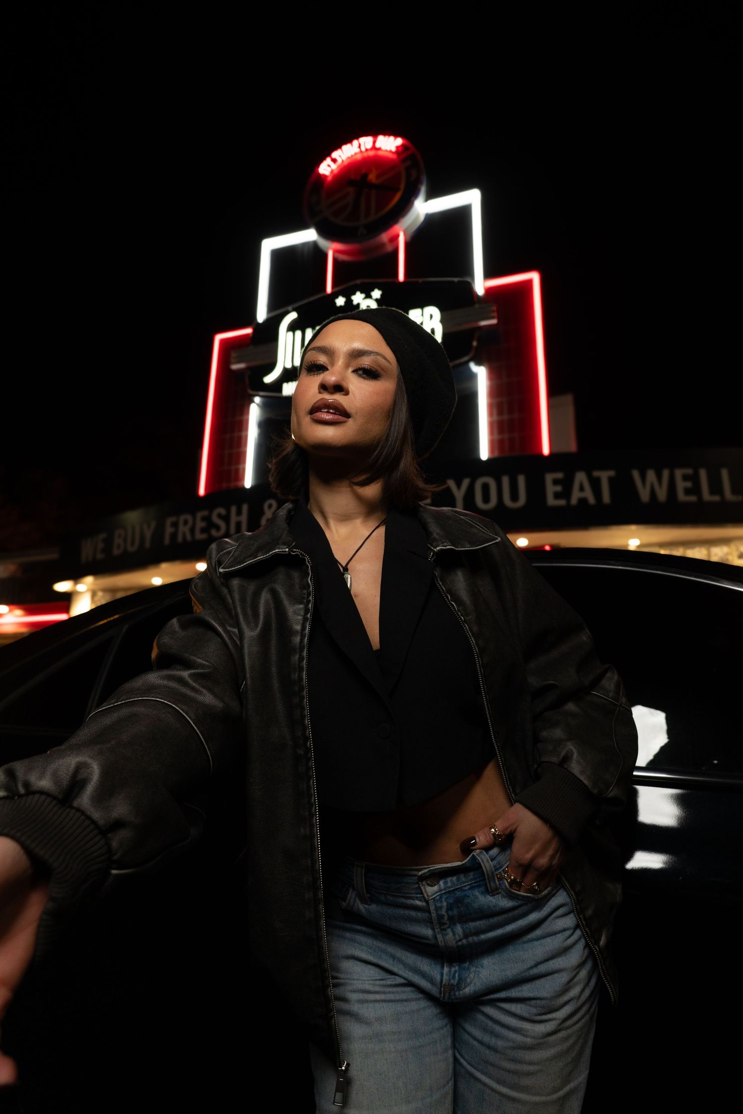 A figure in a black leather jacket and jeans poses dramatically against a dark background with neon signage.