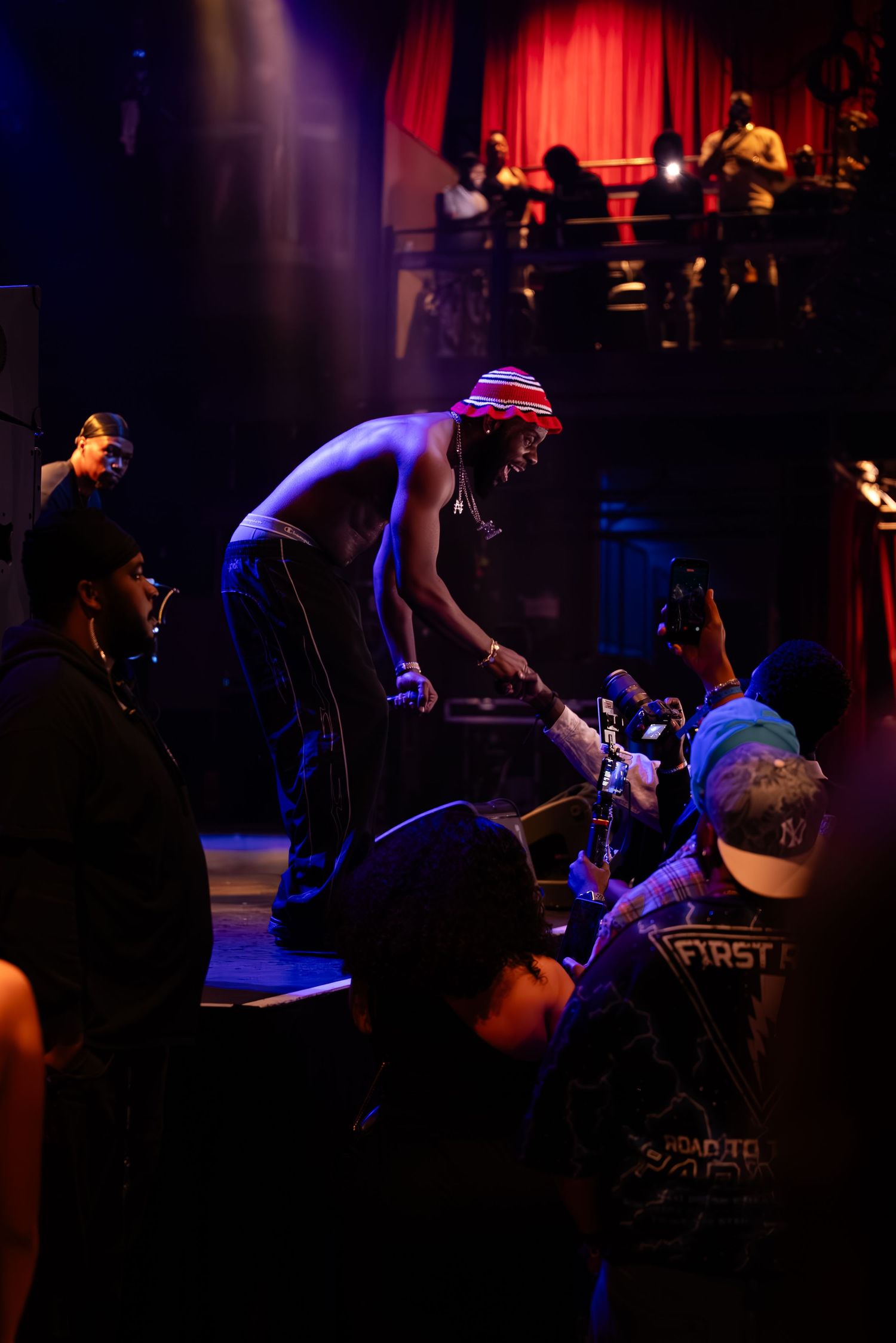 Shirtless performer leans toward crowd at dimly lit concert venue with red curtains and balcony.