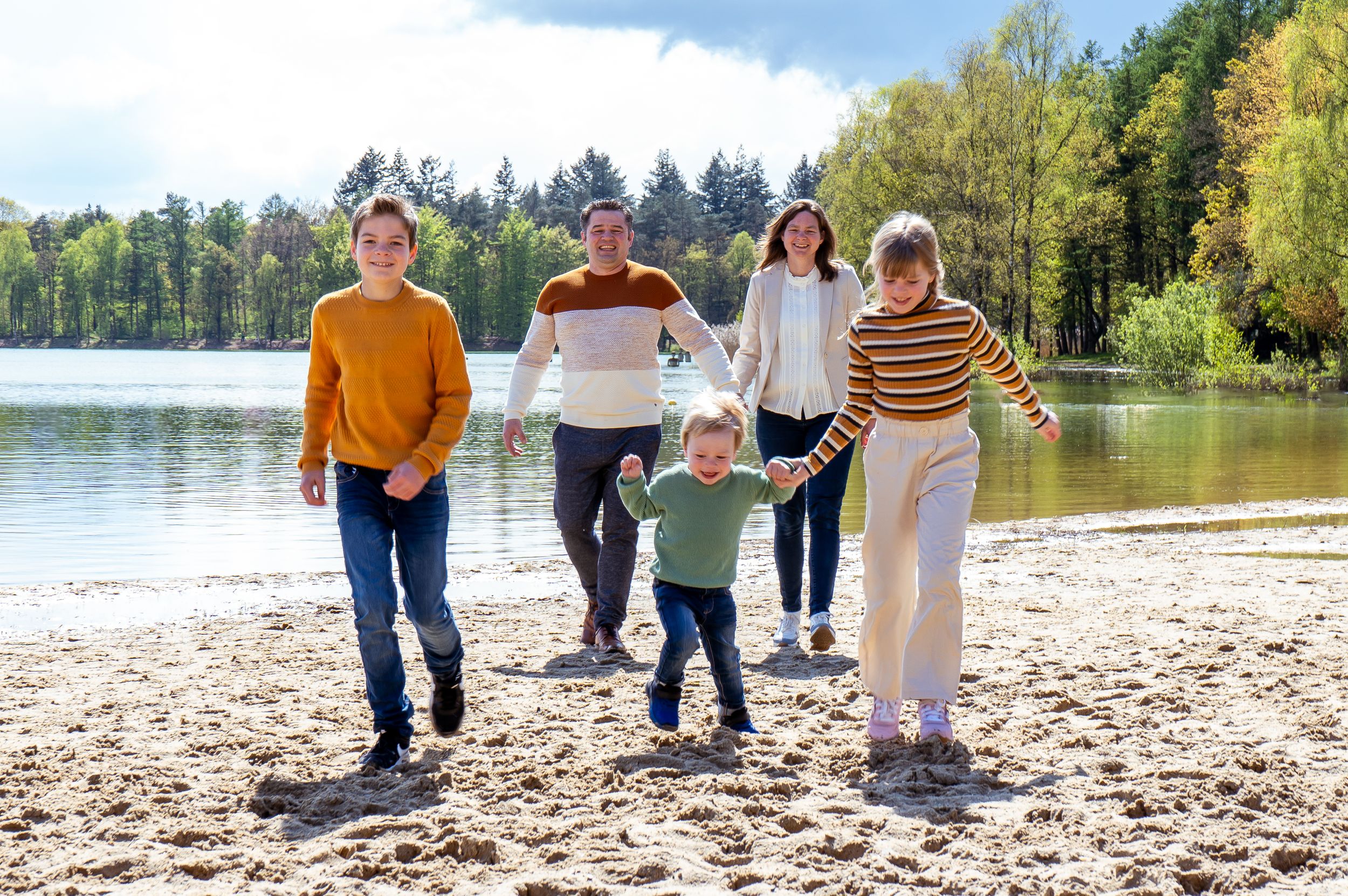 Een gelukkig gezin van vijf wandelt samen over een zandstrand met een meer en bos op de achtergrond.